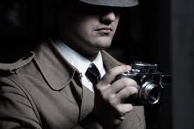 A private investigator, clad in a trench coat and fedora, is discretely taking photographs with a camera. The detective is peeking from behind a wall in a dimly lit alleyway, capturing images of a suspect for a case.